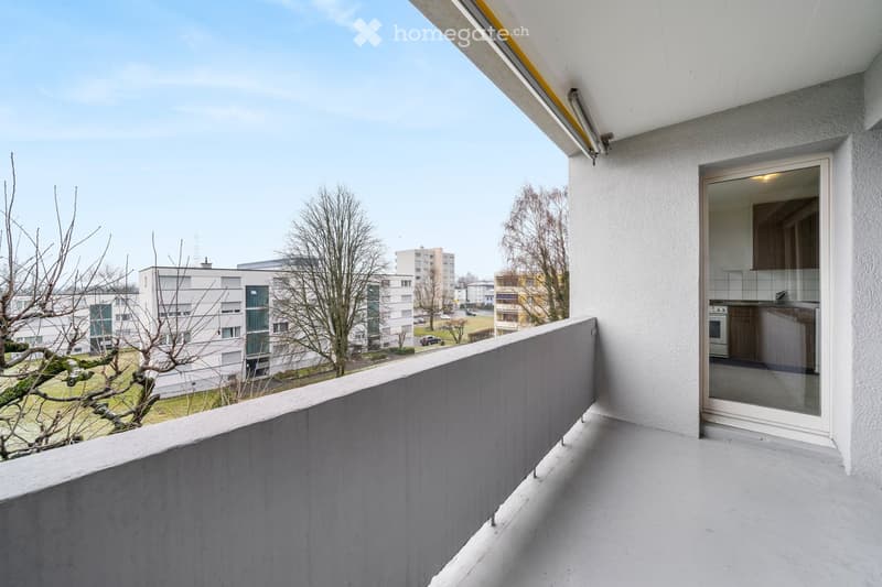 Balcony with a modern design, featuring a grey railing and a view of a residential area with buildings and trees.