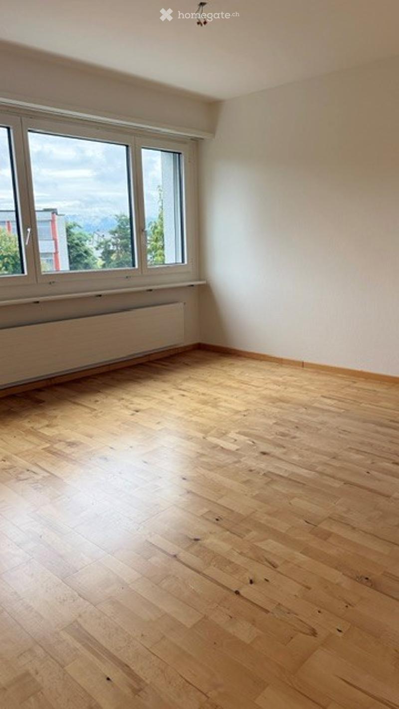 Completely empty room with light wooden flooring and white walls. Large windows provide natural light and a view of the outside.