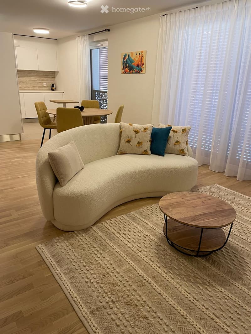 Modern living room with a curved sofa, coffee table, dining area, and large windows with white curtains.