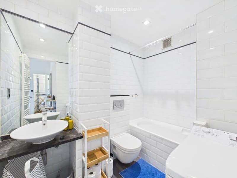 Modern bathroom with white tiles, a sink, toilet, bathtub, and a small shelf holding towels and toiletries.