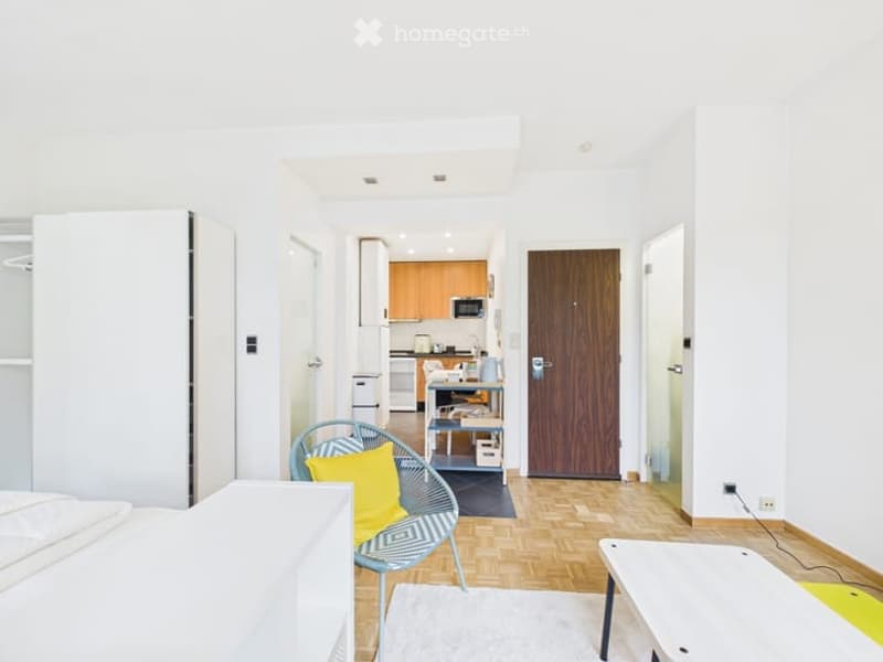 A compact, well-lit studio apartment with a bed, a modern chair, a small table, and an open kitchen area featuring cabinets, a microwave, and a refrigerator.