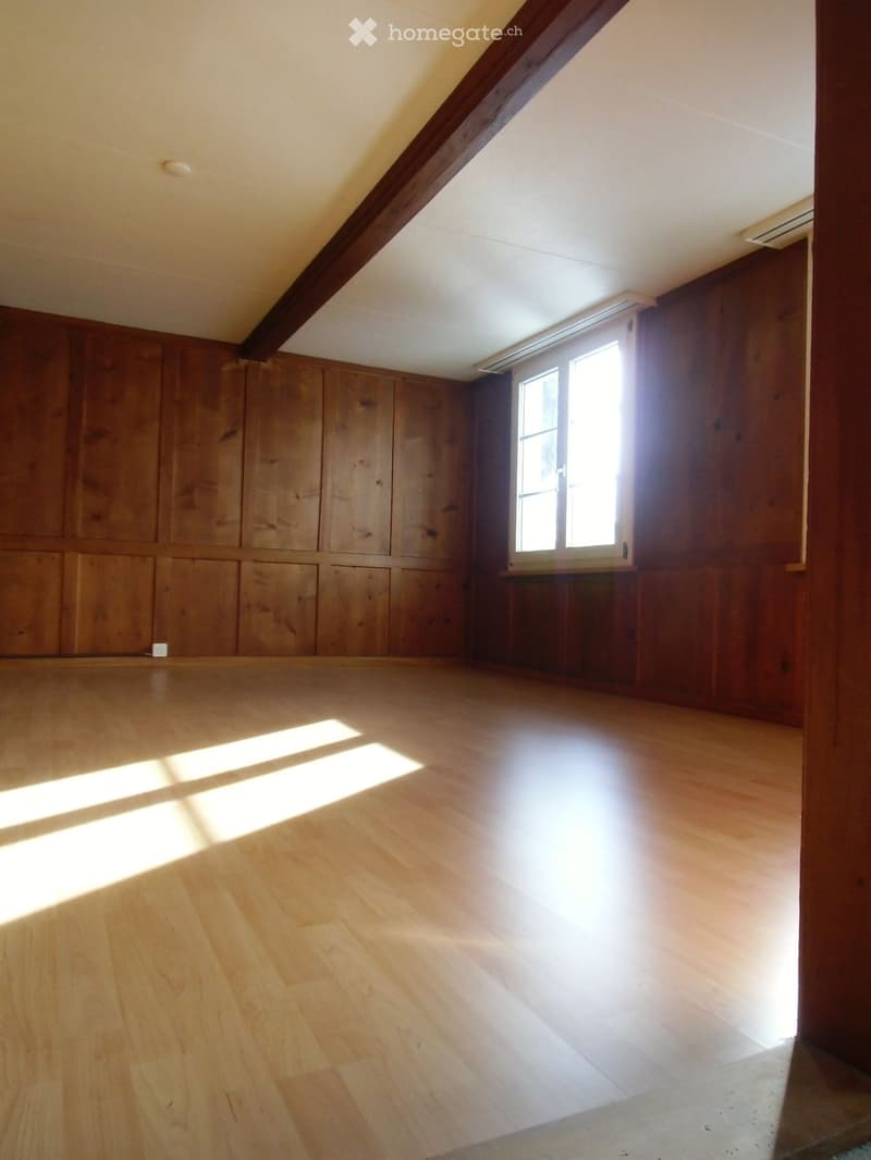 Empty room with wooden walls and floor, large window allowing natural light, exposed wooden beam on the ceiling.