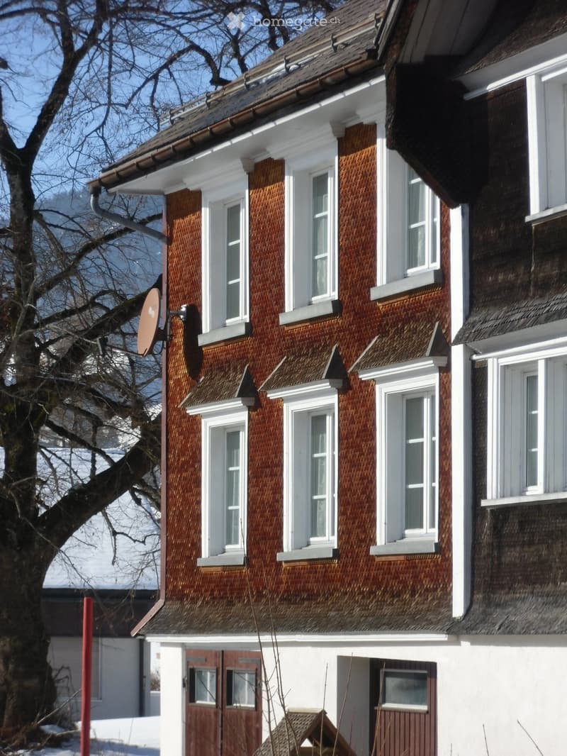 Two-story house with brown tiled exterior, white trim around windows and roofline, multiple windows, and a satellite dish mounted on the wall.