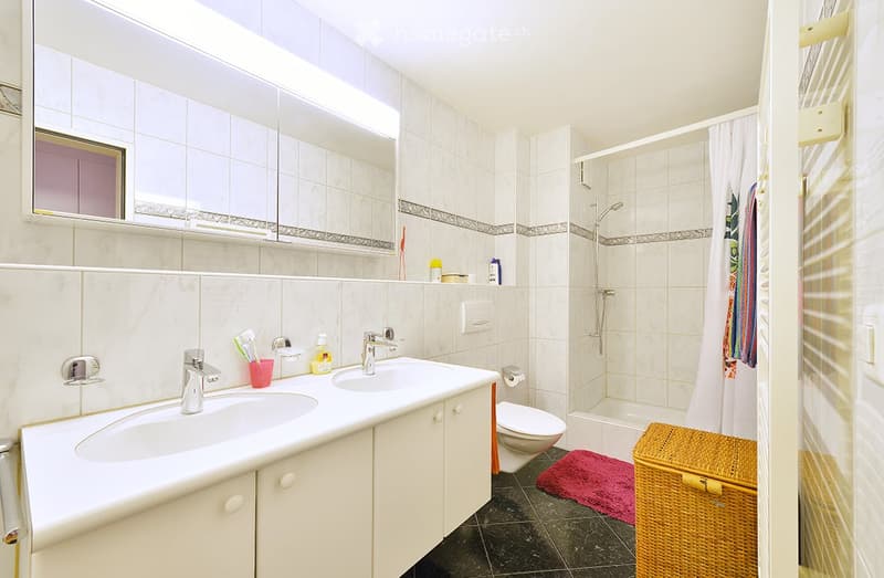 Modern bathroom with white tiles, double sink, large mirror, shower, and a wicker basket on the floor.