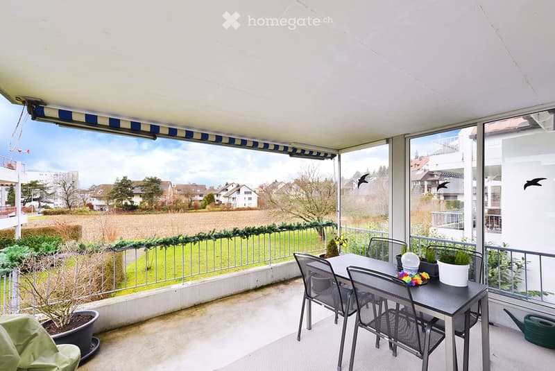 Balcony with a table, chairs, and potted plants, offering a view of a green landscape and houses.