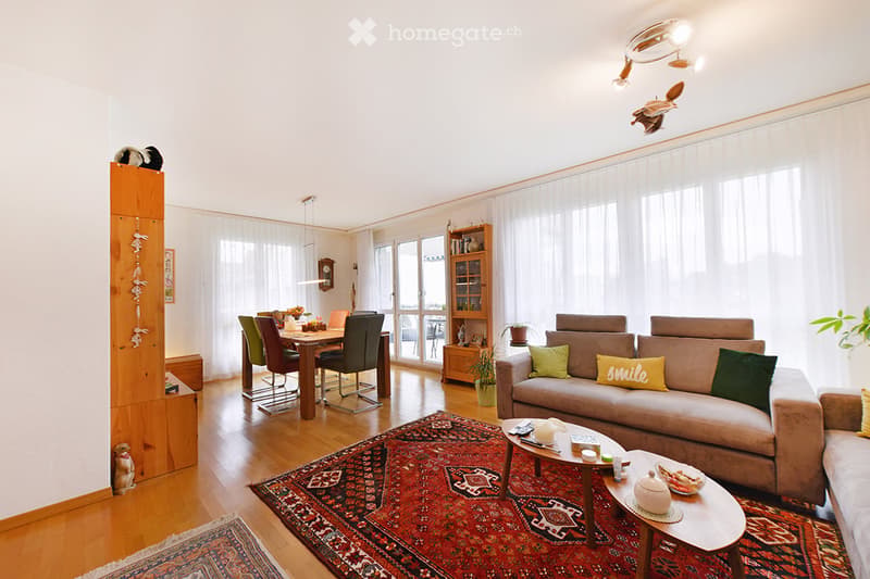 Bright, open living and dining area with wooden flooring, large windows, a beige sofa with colorful cushions, a coffee table, a dining table with chairs, a bookshelf, and decorative items.