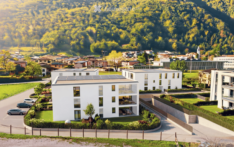 Modern apartment complex with solar panels on the roof, situated on a raised ground floor, surrounded by greenery