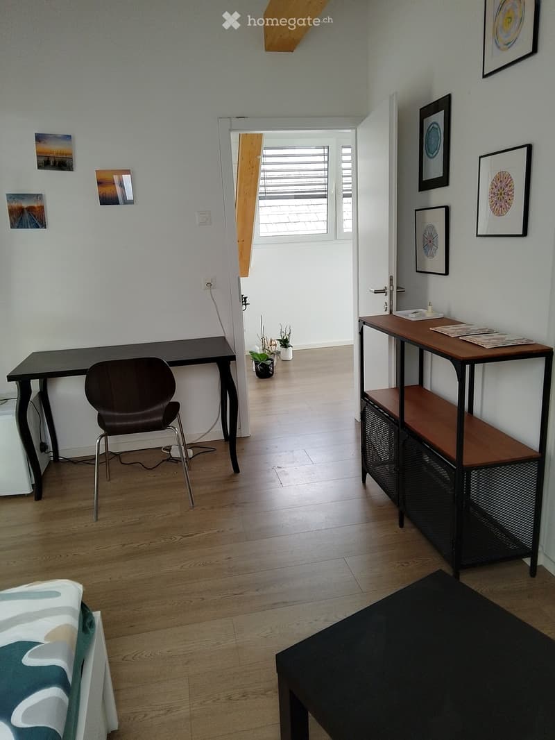 A modern, well-lit room with wooden flooring, a sofa, a desk with a chair, a small table with a shelf, and framed artwork on the walls.