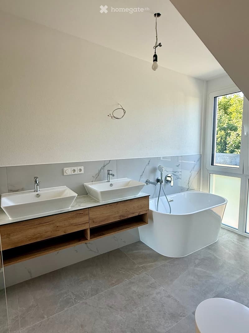 Modern bathroom with two white sinks on a wooden vanity, a freestanding bathtub, and a large window offering a view of greenery.