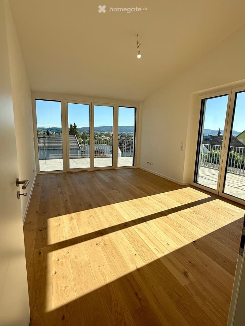 Empty room with wooden flooring, large windows, and sliding doors leading to a balcony. Natural light fills the space.