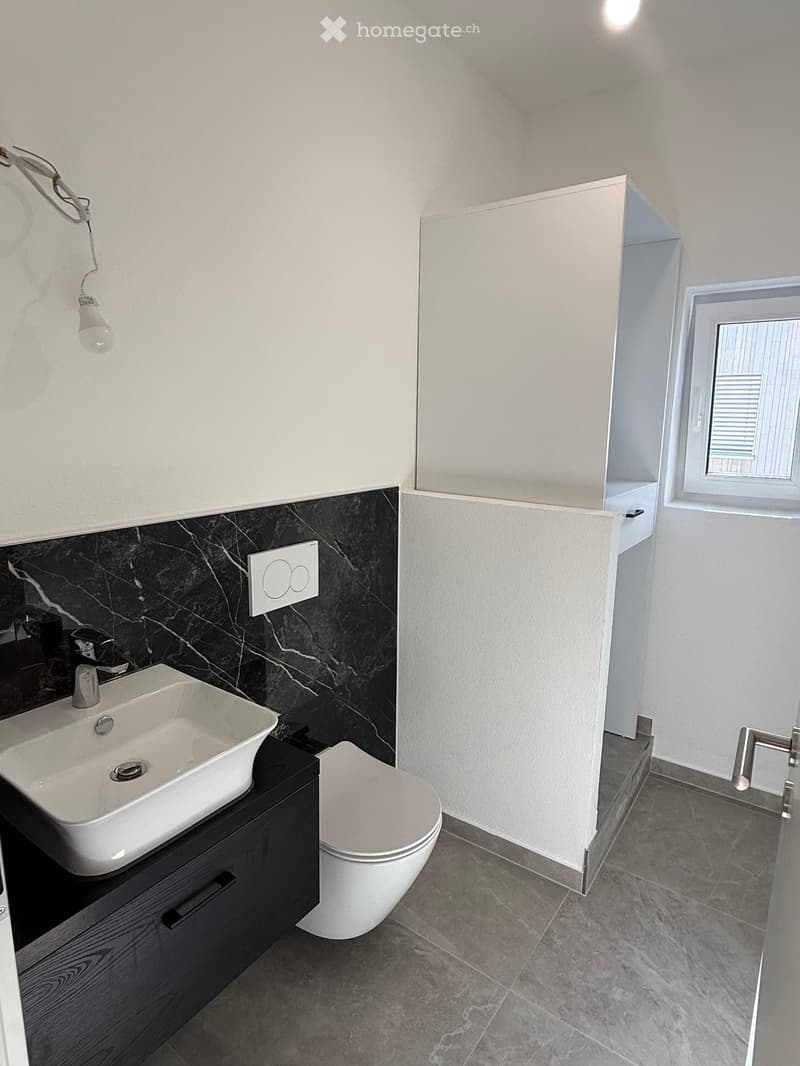 Modern bathroom with a white sink, chrome faucet, toilet, and dark wooden cabinet. The walls feature a black marble accent above the sink, and the floor is tiled in gray.
