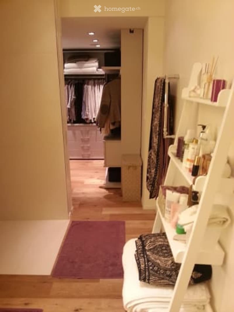 Bathroom with wooden floor, white walls, shelf with towels and toiletries, and an open closet with clothes visible.