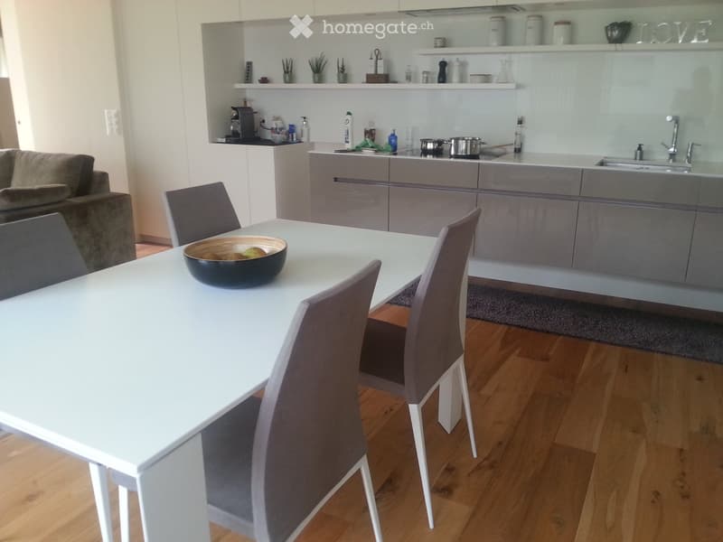 Modern kitchen and dining area with grey cabinets, white dining table, grey chairs, and a sofa in the background.
