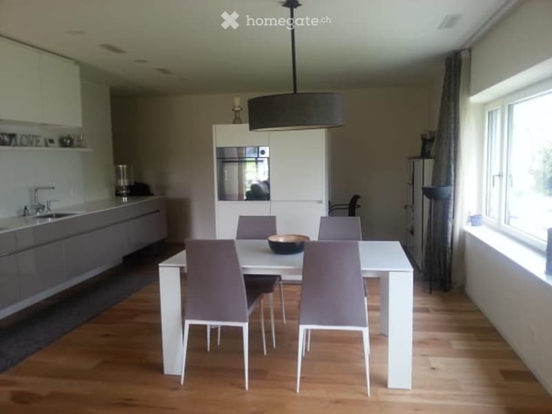 Modern dining area with a white dining table and grey chairs, integrated kitchen with cabinets and a sink, pendant light fixture, and large windows with curtains.
