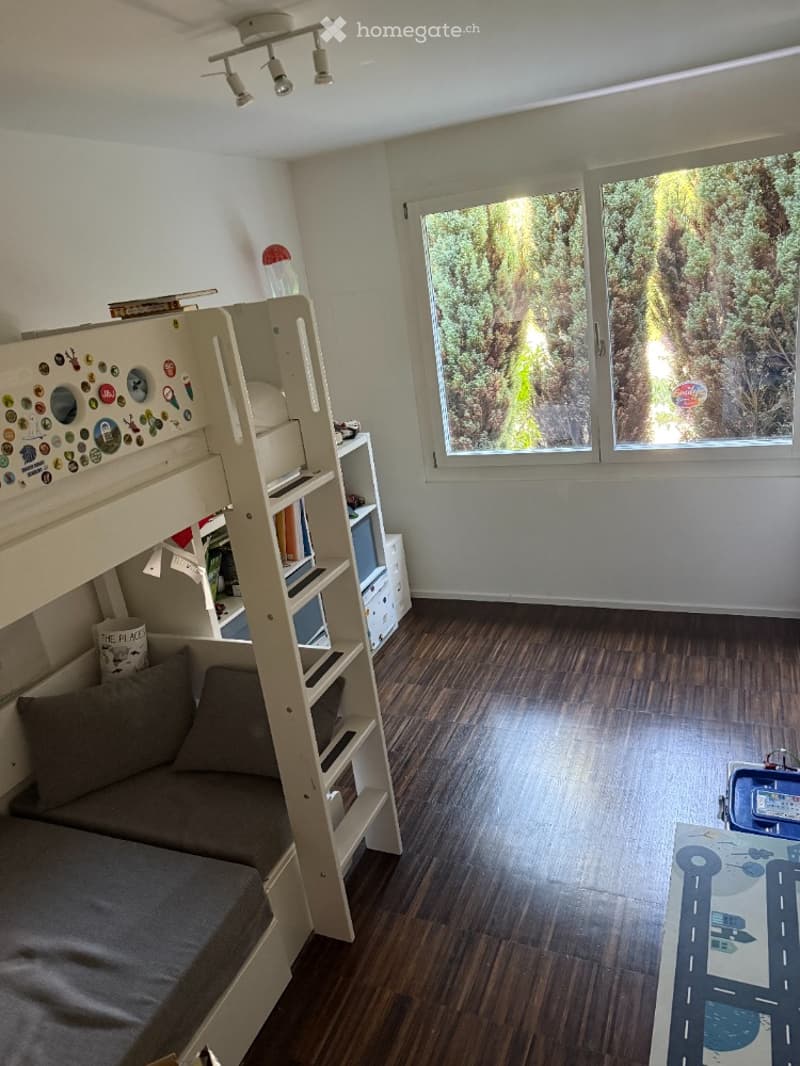 Cozy bedroom with a bunk bed, shelves, a small table, and a view of greenery outside.