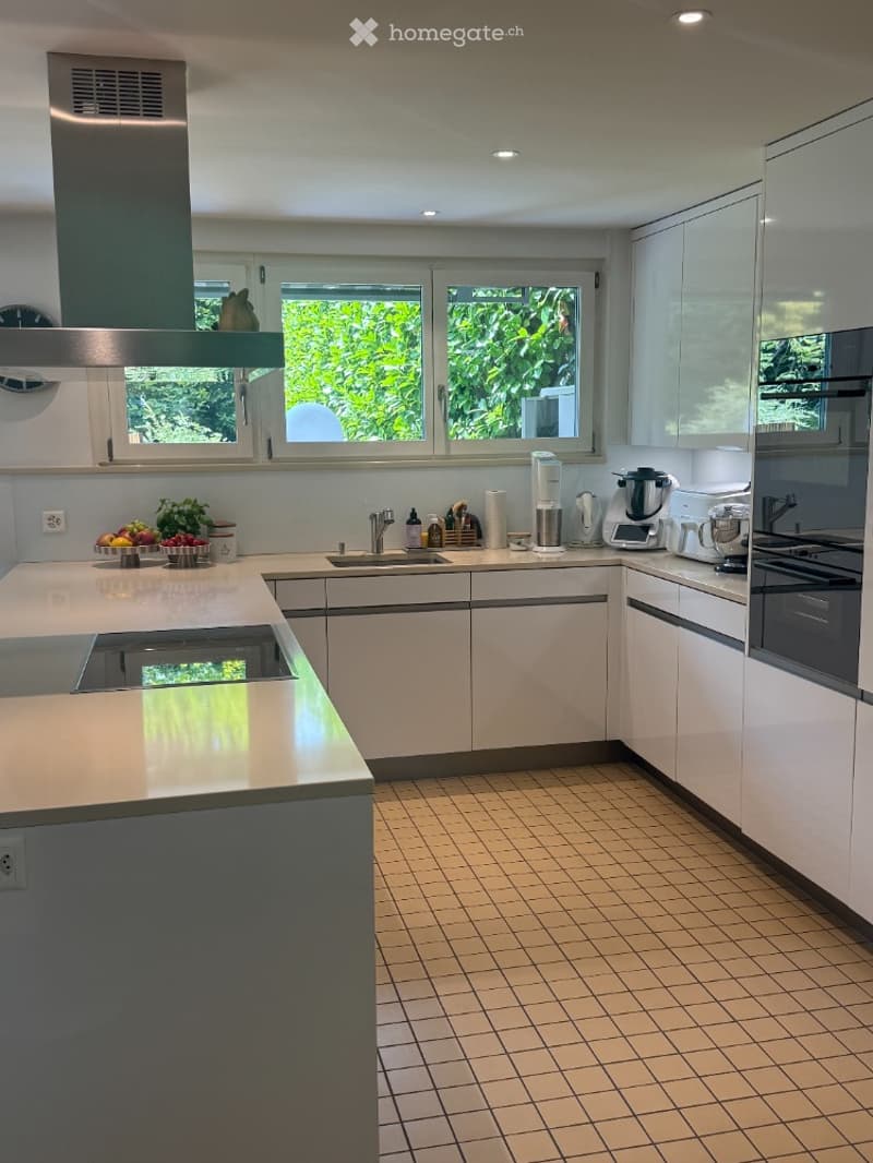 Modern kitchen with white cabinets, light-colored countertops, and a tiled floor. Features include a sink, faucet, oven, blender, coffee maker, and a fruit bowl on the counter.
