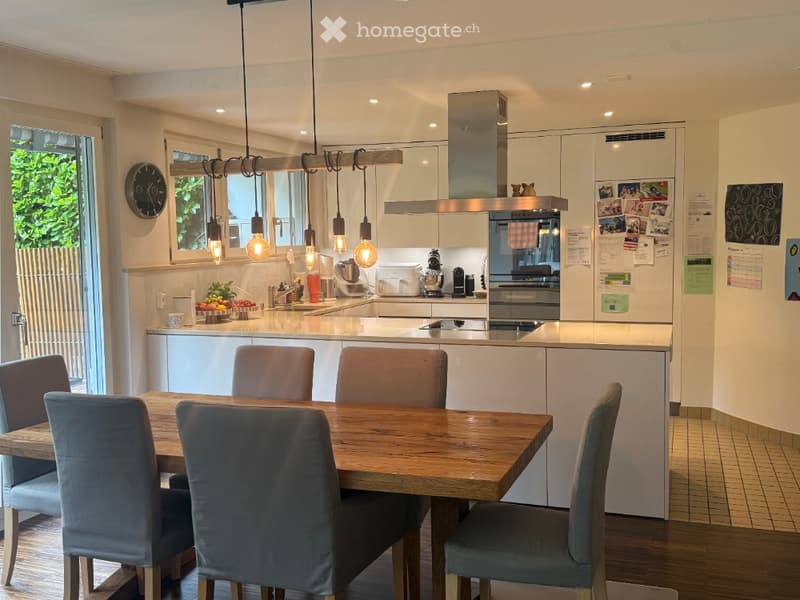 Modern kitchen with white cabinets, a central island, a dining table with six chairs, pendant lights, and various appliances including a refrigerator, oven, and blender. A fruit bowl is on the counter, and a clock is mounted on the wall.