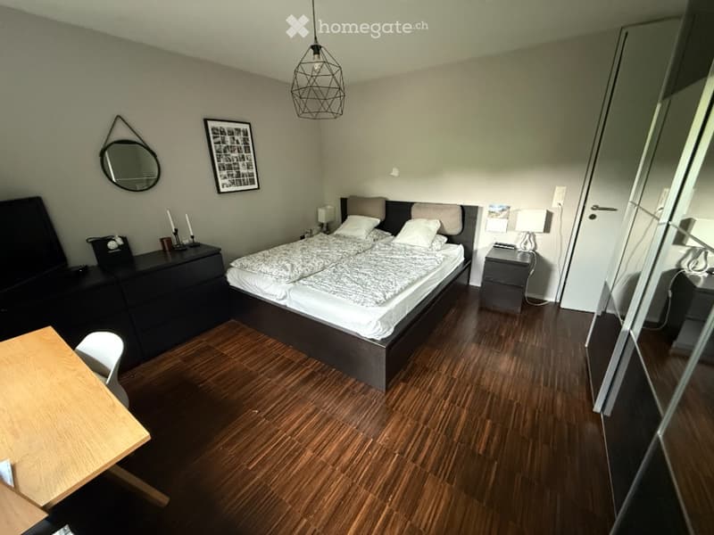 A modern bedroom with a double bed, nightstands, a mirror, a chair, and a small table. The room features wooden flooring and a hanging pendant light.