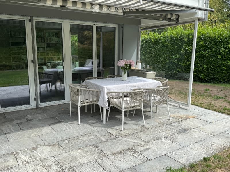 Outdoor patio with a dining set, white wicker chairs, and a table covered with a white tablecloth. The area is covered by a striped awning and features large glass doors leading to the interior.