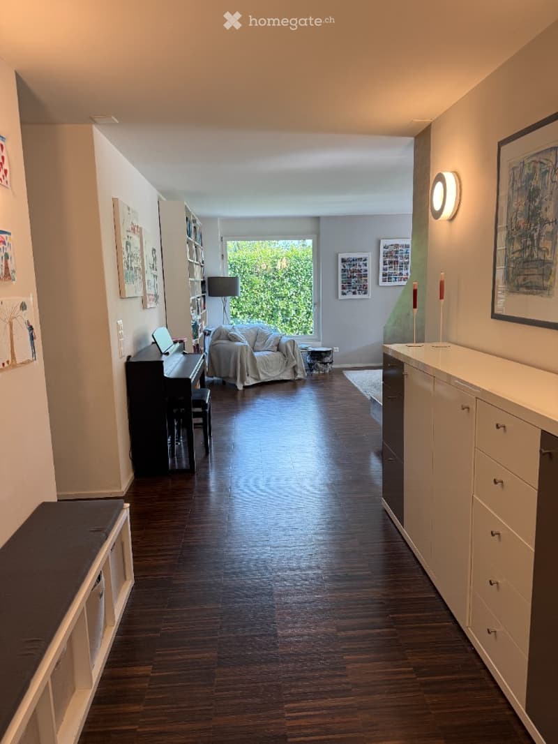 A spacious, well-lit living room with hardwood flooring, a sofa, a piano, a laptop on a table, a bookshelf, and a cabinet with drawers.