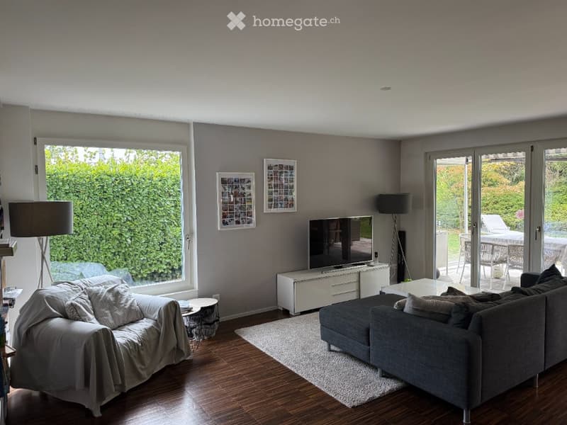 Modern living room with grey walls, large windows, and a view of a garden. Features a grey sectional sofa, coffee table, TV stand, and floor lamp.