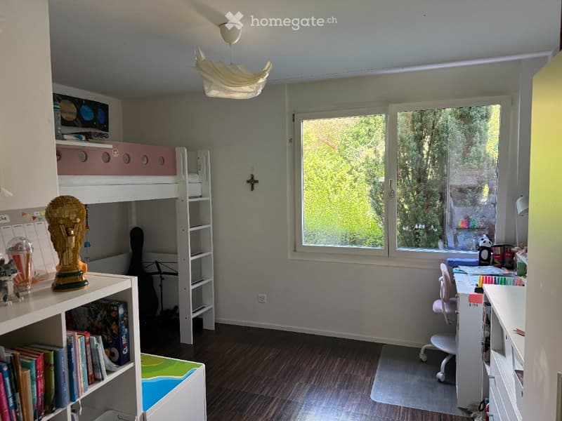 A well-lit bedroom with a bunk bed, a desk with a chair, a shelf filled with books, and a guitar leaning against the wall. A large window offers a view of greenery outside.
