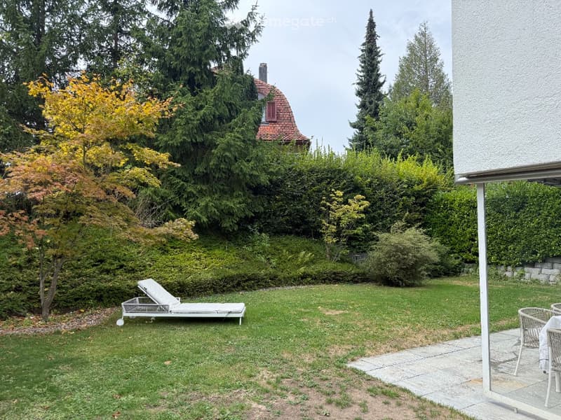 Well-maintained garden with a sun lounger, outdoor seating, and a variety of trees and shrubs.