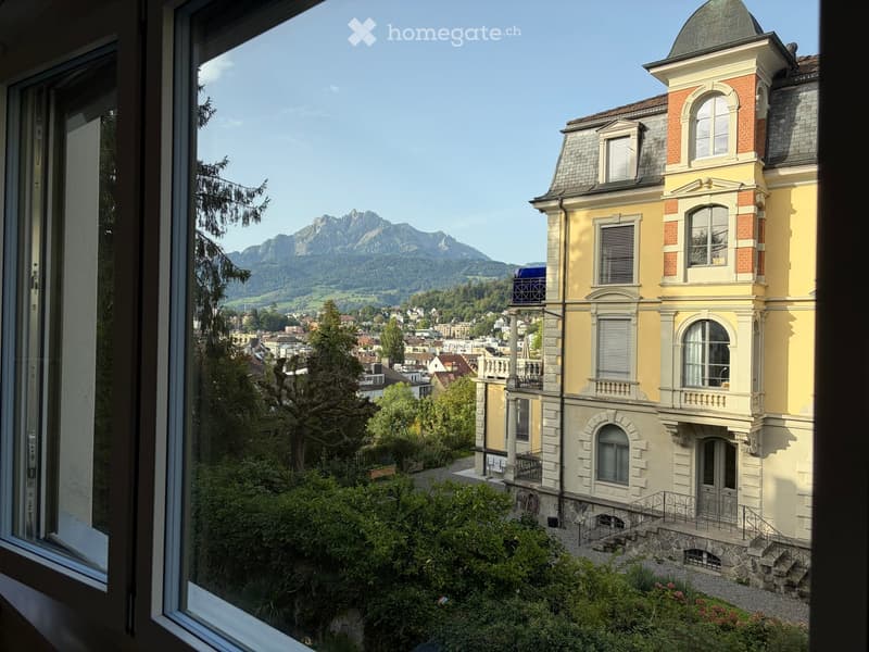 View from a window showing a large, yellow and white building with multiple stories and a dome-shaped roof, surrounded by greenery and trees, with a scenic mountain view in the background.