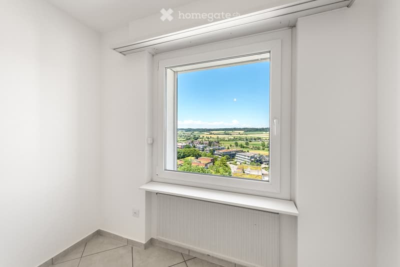Empty room with white walls, tiled floor, large window offering a view of a suburban landscape, radiator below the window, and a small shelf on the windowsill.