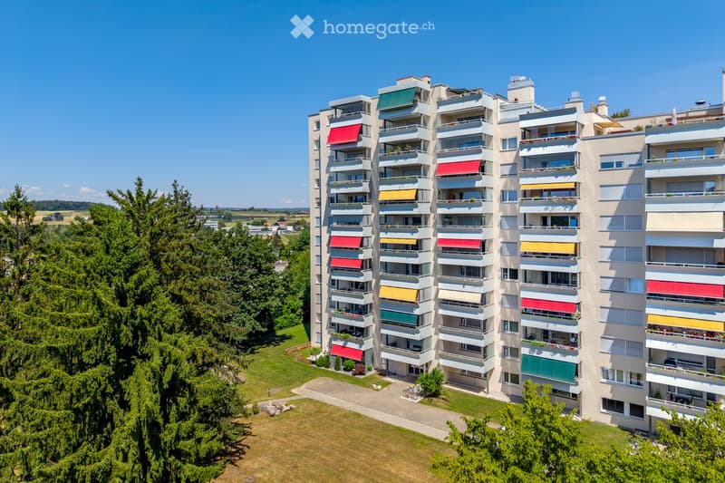 A modern multi-story apartment building with balconies, colorful awnings, and surrounding greenery.