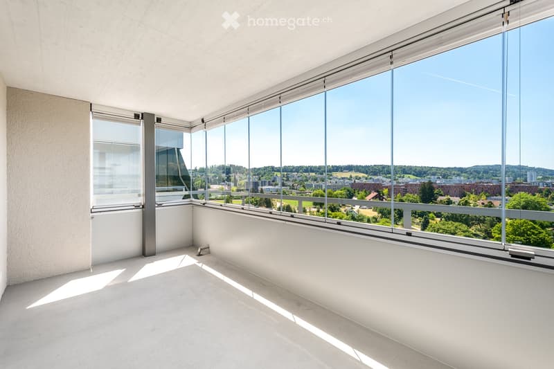 An empty room with large floor-to-ceiling windows offering a panoramic view of a cityscape and greenery. The room features a smooth concrete floor and white walls.