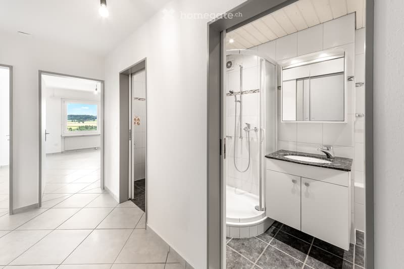 Bright hallway with white tiled floors and walls, leading to a modern bathroom with a shower stall, sink, and mirrored cabinet.