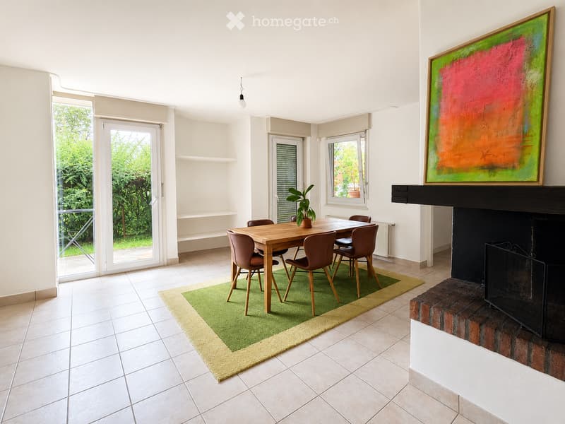 Bright dining room with a wooden dining table, six chairs, a green area rug, a fireplace with a painting above it, and a potted plant on the table. Large windows and sliding glass doors lead to a garden.