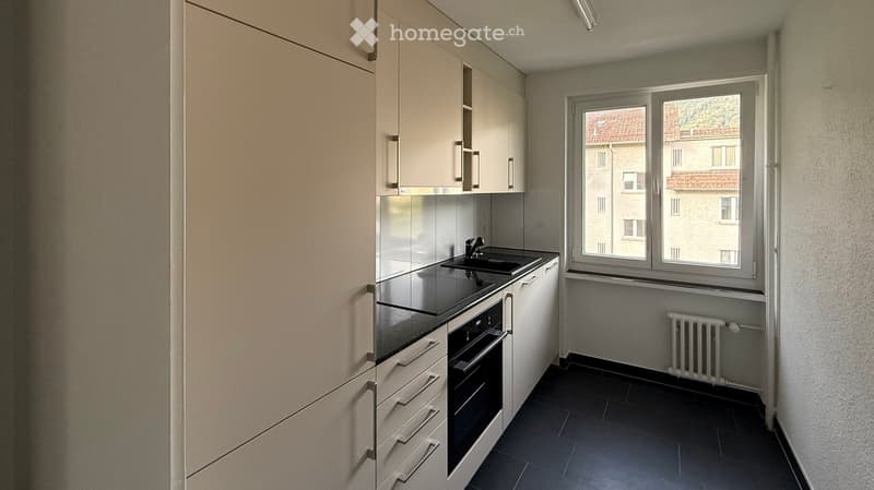 Modern kitchen with white cabinets, black countertop, sink, oven, and radiator. Features a large window with a view of neighboring buildings.