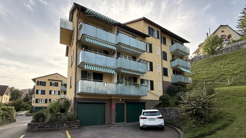 Multi-story apartment building with balconies, green garage doors, and a parked car in front.