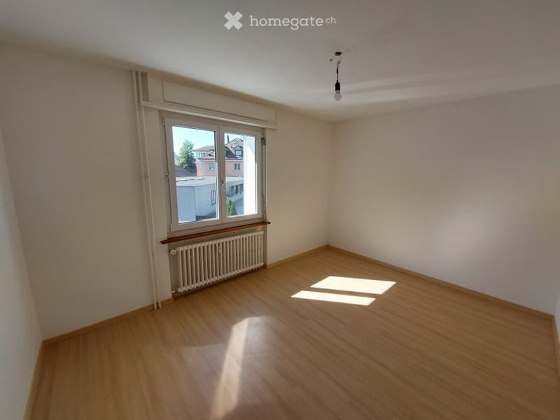 Empty room with wooden flooring, white walls, and a large window with a radiator below it. No furniture or equipment is present.