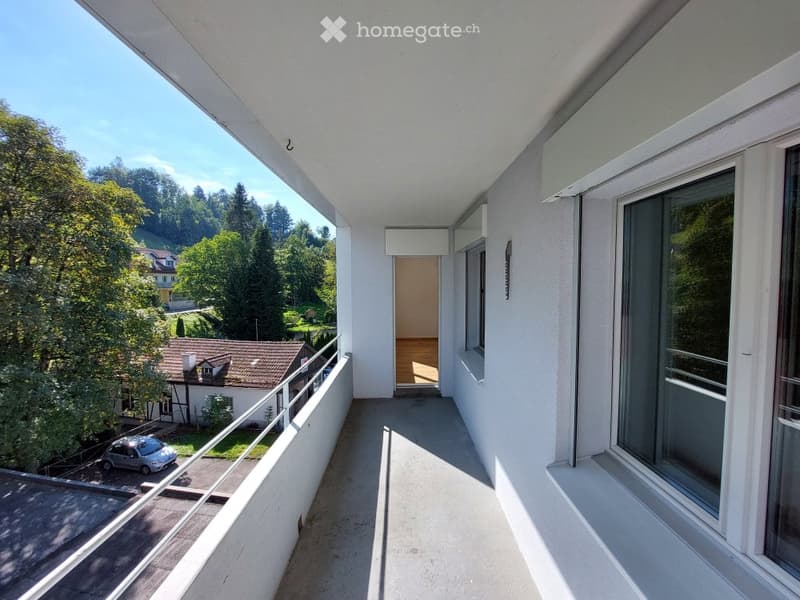 Balcony with a view of a green, hilly landscape, featuring a metal railing, large windows, and a door leading to an interior space.