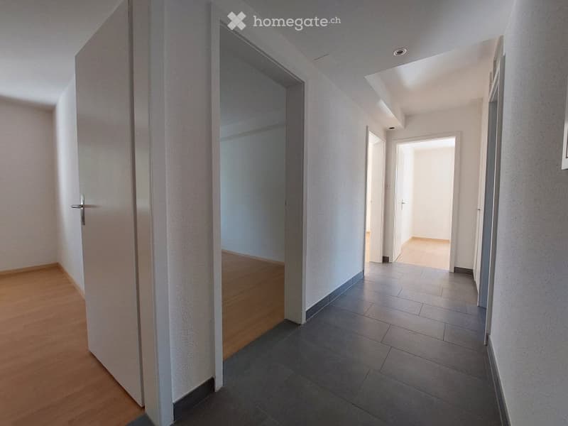 Empty hallway with wooden flooring on one side and tiled flooring on the other, featuring several doors and a bright, open layout.