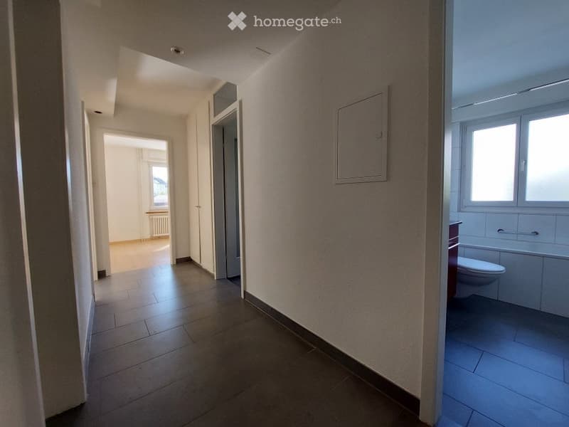 Empty hallway with tiled floor, white walls, and doors leading to other rooms, including a bathroom with a toilet and bathtub.