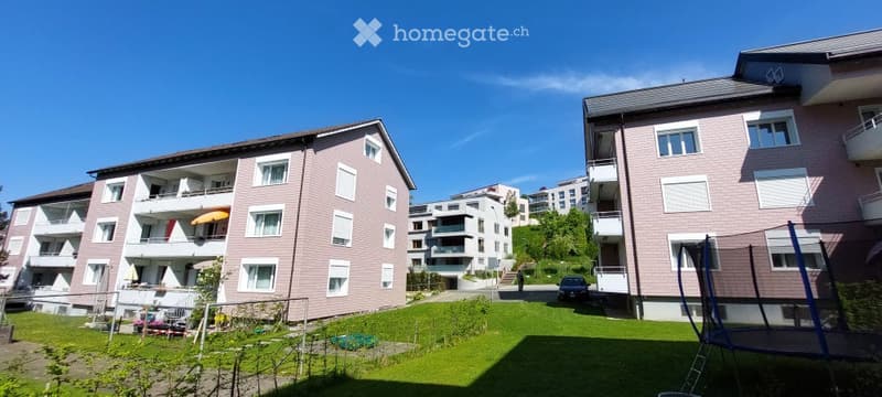 A multi-story residential building with pink facades, white-framed windows, and balconies. The building is surrounded by a well-maintained lawn and includes a small playground area with a trampoline and swings.