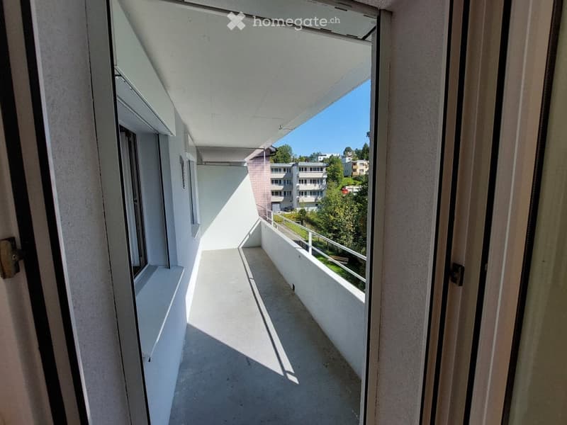 Modern balcony with white walls, concrete floor, and metal railing, overlooking a residential area with trees and buildings.