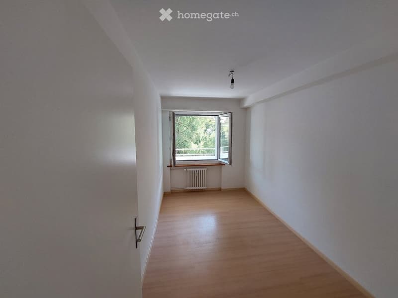 Empty room with wooden flooring, white walls, and a window with open shutters. A radiator is visible beneath the window, and a single ceiling light fixture is present.