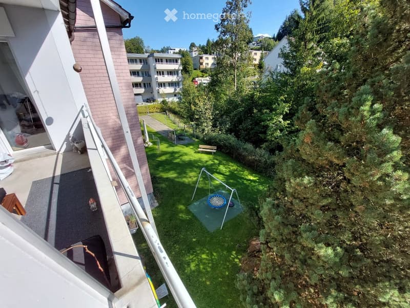 A sunny, green backyard with a swing set, a small bench, and a grassy area surrounded by trees and neighboring buildings.