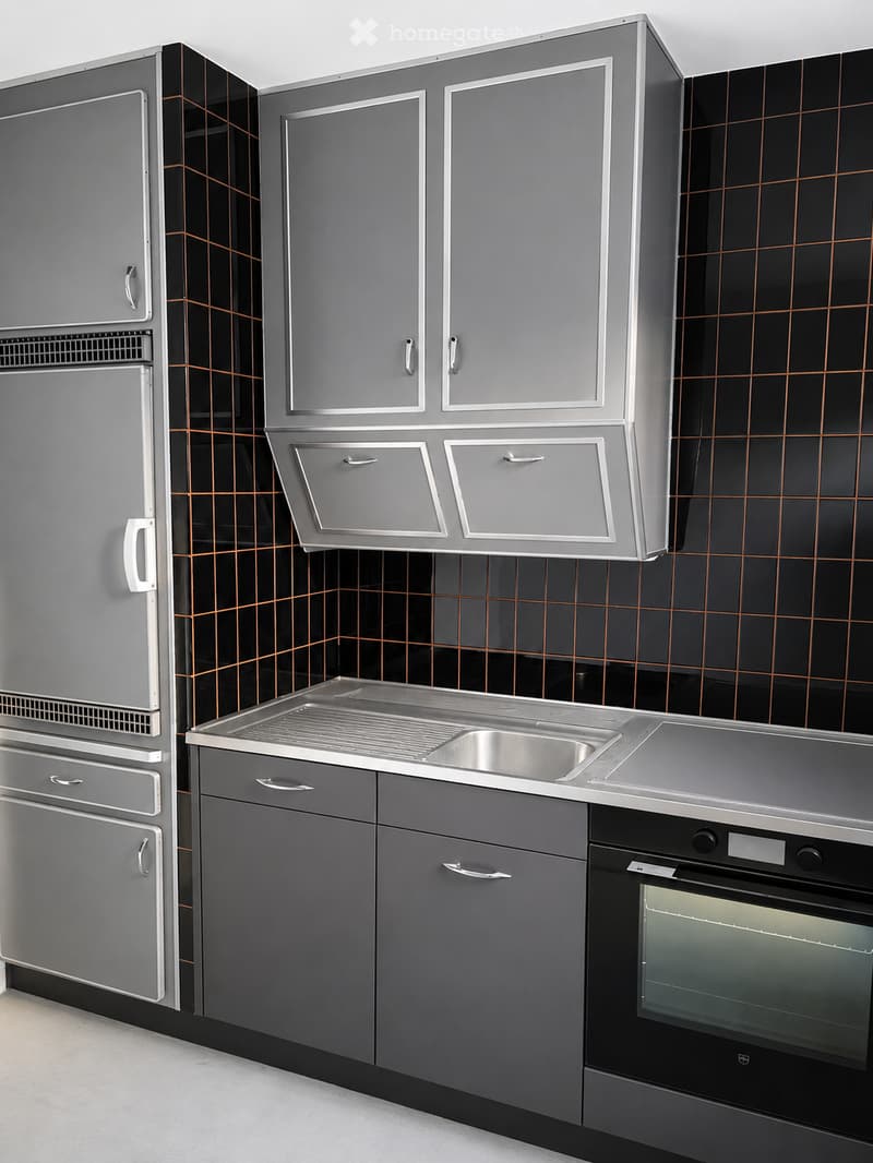 Modern kitchen with grey cabinets, stainless steel sink, refrigerator, and oven. Black tiled backsplash with copper grid pattern.