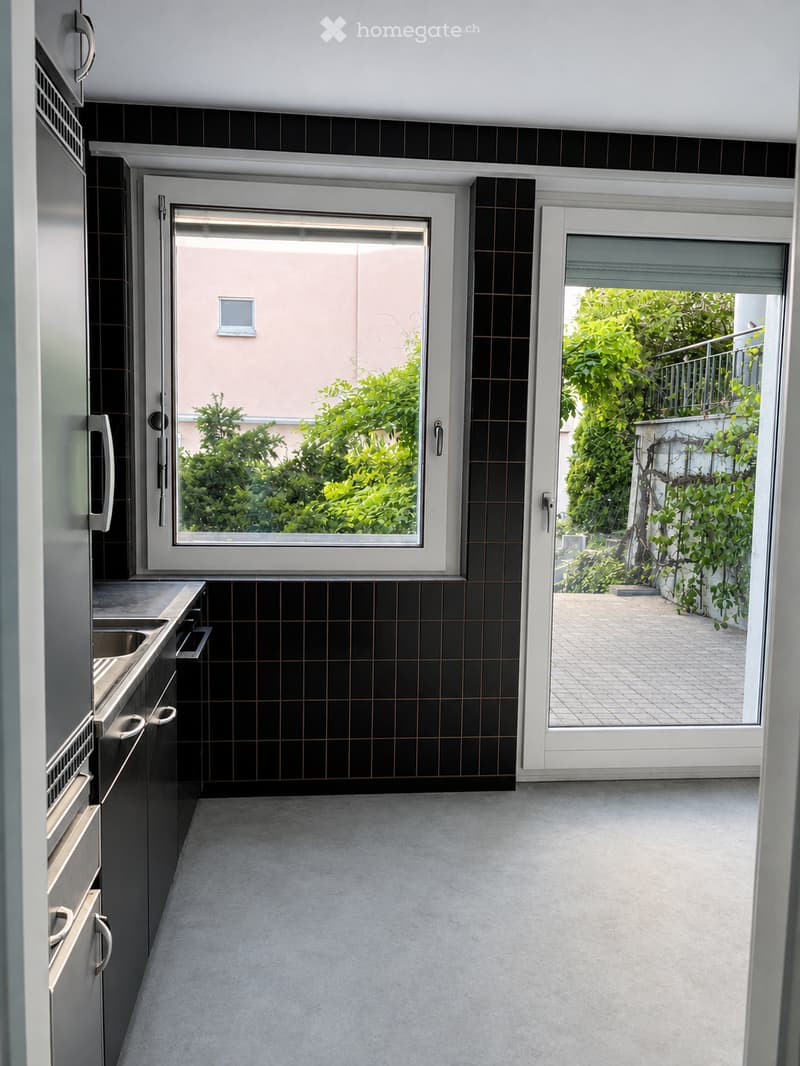 Modern kitchen with black tiled walls, grey cabinets, and a large window overlooking greenery. Features a sink and a sliding door leading to a patio.