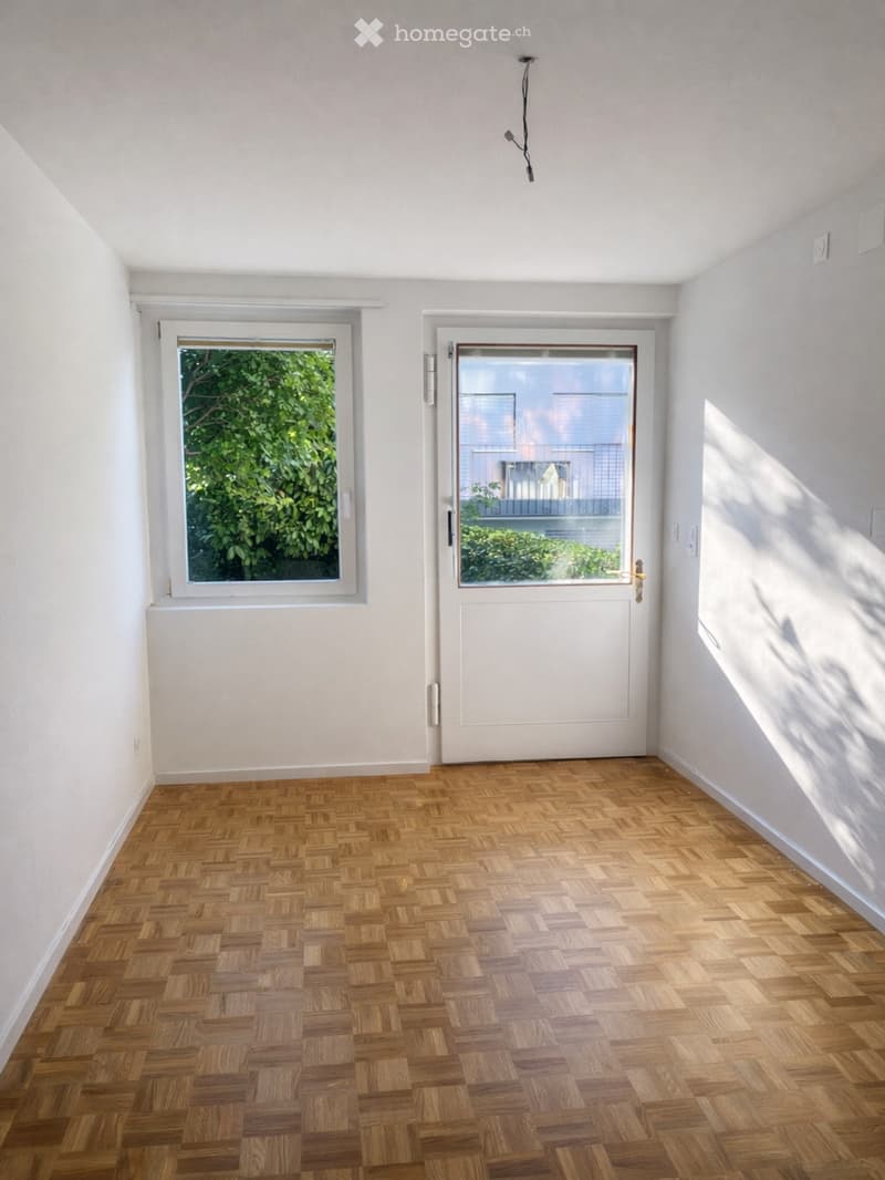 Empty room with wooden herringbone flooring, white walls, and large windows with white frames. A white door with a glass panel leads to an outdoor area.