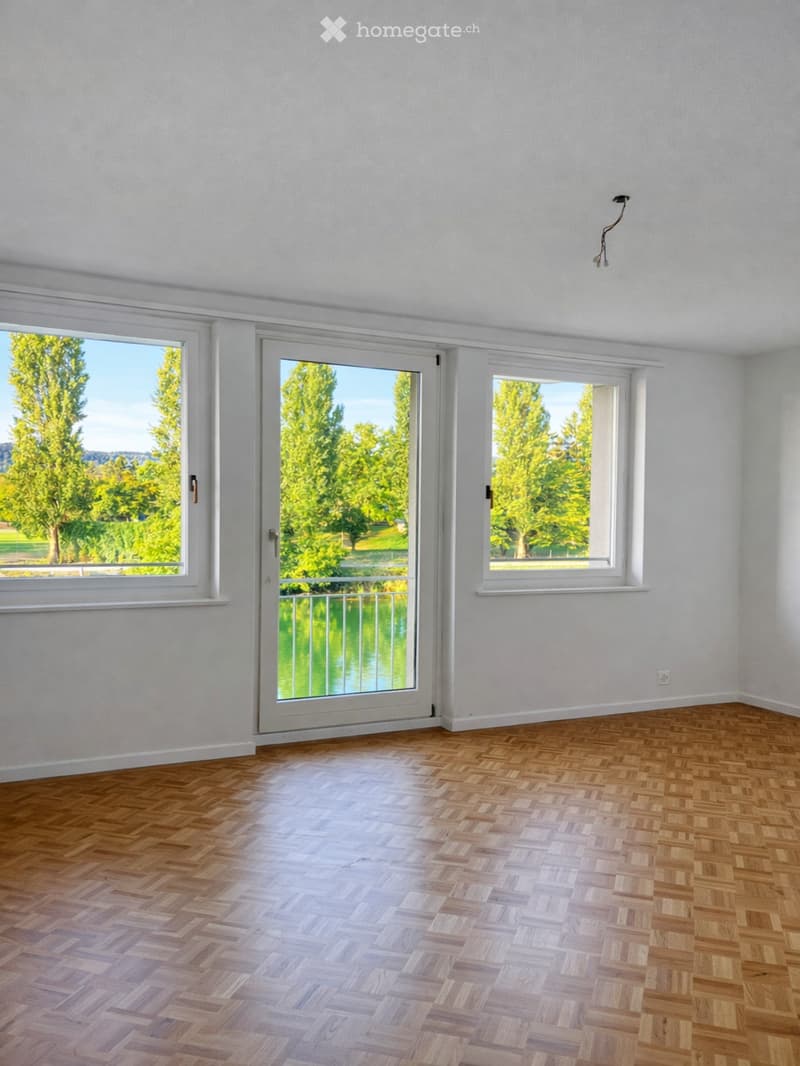 Empty room with wooden herringbone flooring, white walls, large windows, and a sliding glass door leading to a balcony with a view of greenery.
