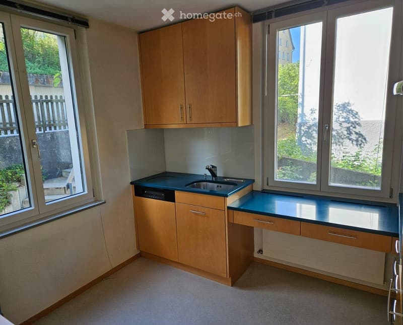 Modern kitchen with wooden cabinets, dark countertops, and a sink with a faucet. Large windows provide natural light and a view of greenery outside.