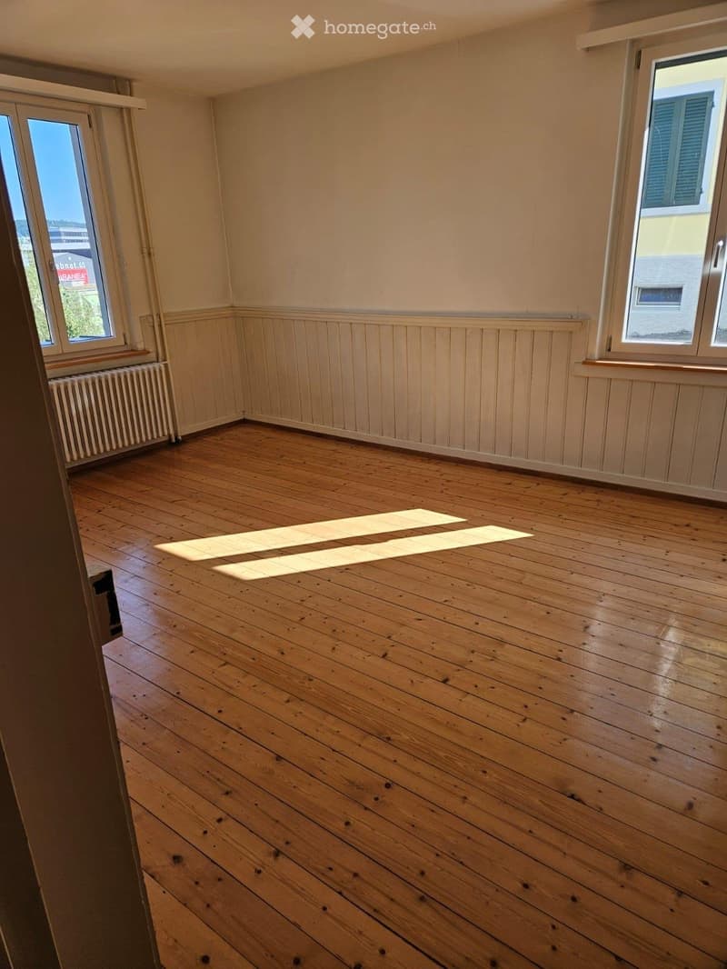Empty room with wooden flooring, white walls, and large windows allowing natural light to enter. The room features a radiator on one wall and a visible baseboard trim.