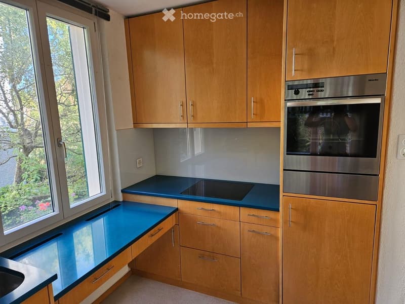 Modern kitchen with light wooden cabinets, blue countertops, and a built-in oven. Large window offers a view of greenery outside.